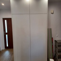 Adrian Świerczyński - Biała szafa wnękowa z czterema drzwiami i oświetleniem punktowym w suficie, widoczne narzędzia i akcesoria montażowe obok szafy.