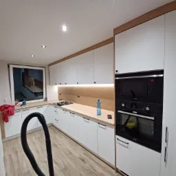 Nowoczesna kuchnia z białymi frontami, drewnianymi blatami i czarnym sprzętem AGD Amica, widoczny wąż od odkurzacza sugerujący sprzątanie po montażu.