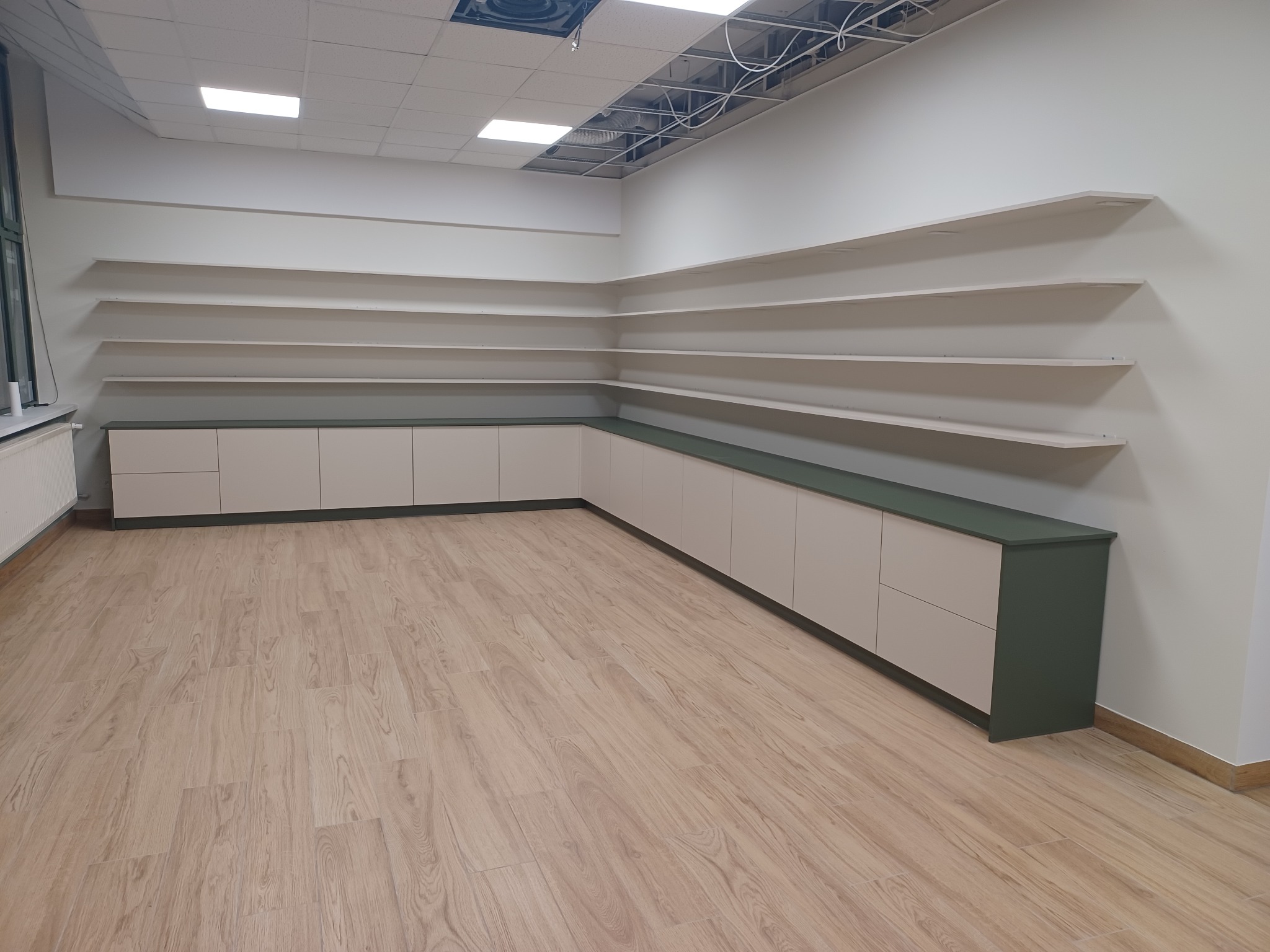 Jasne szafki na wymiar z ciemnozielonym blatem i otwartymi półkami w biurze, widok na podłogę z paneli.