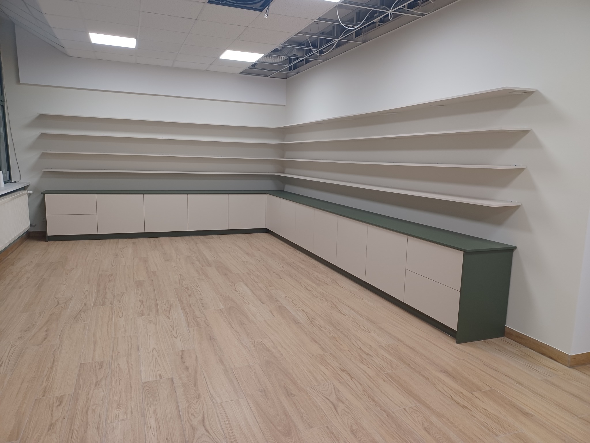 Długa szafka z beżowymi frontami i ciemnozielonym blatem, ciągnąca się wzdłuż ściany, z rzędem półek nad nią, na jasnej podłodze imitującej drewno, w pomieszczeniu z częściowo odsłoniętym sufitem.