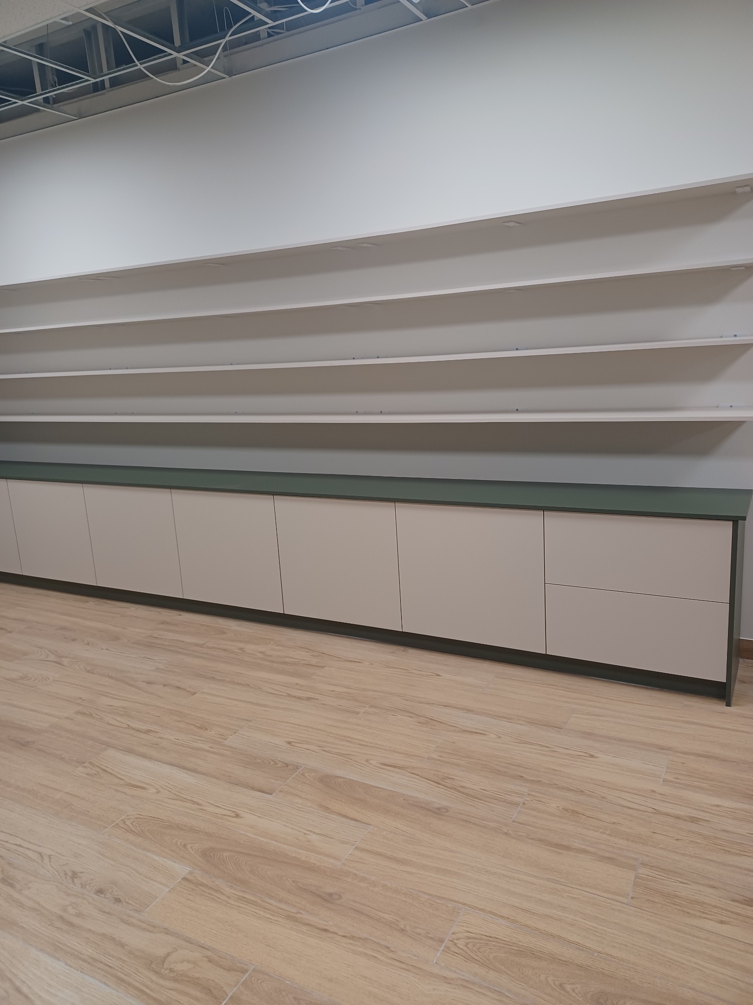 Długi rząd szafek z jasnymi frontami i zielonym blatem, zamontowany wzdłuż ściany z półkami, na podłodze panele imitujące drewno. Widoczne elementy sufitu podwieszanego.