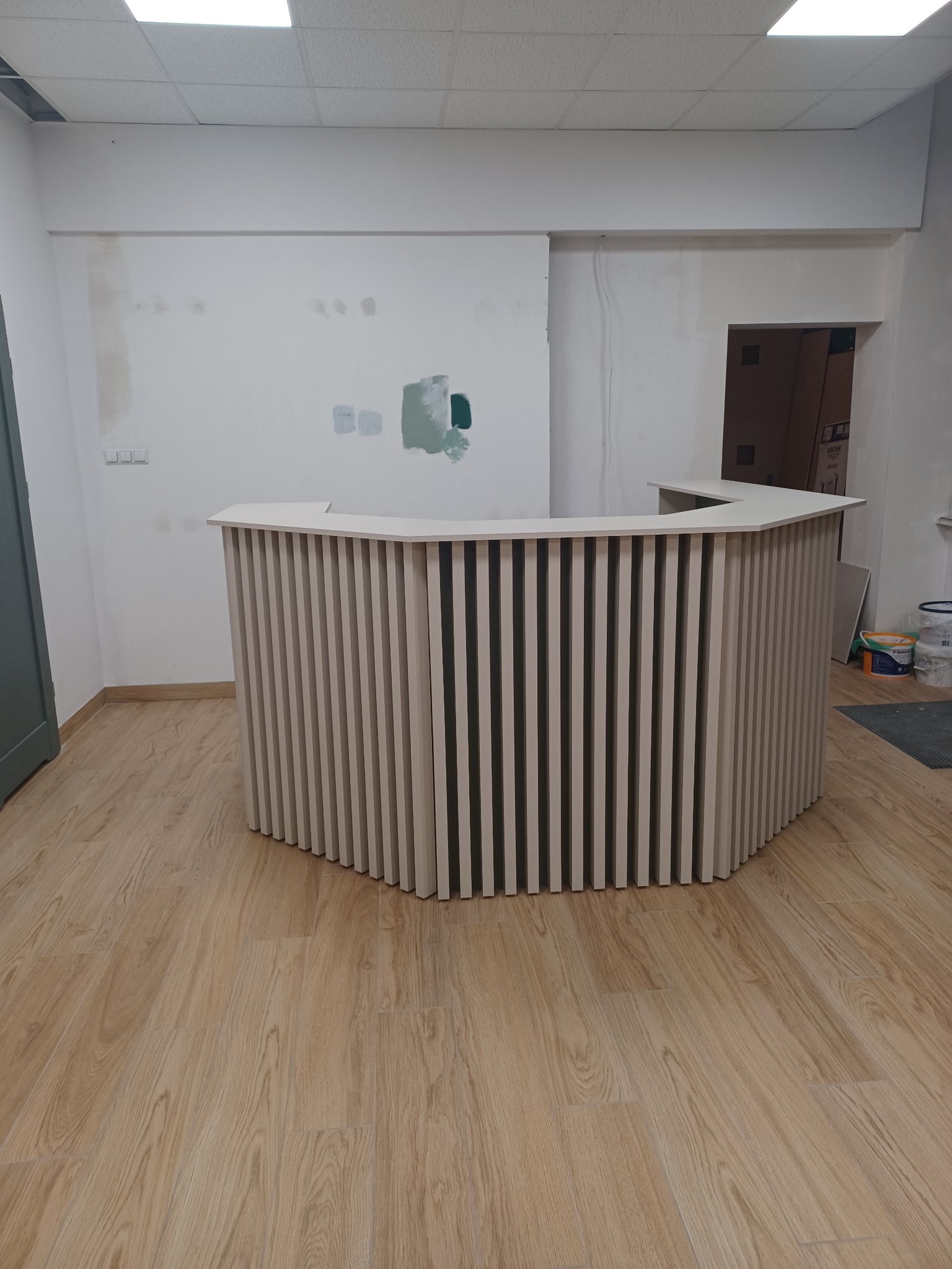 Nowoczesna lada recepcyjna z pionowych listew w jasnym odcieniu, widoczna w surowym wnętrzu przed wykończeniem, z plamami farby na ścianie i drewnianą podłogą.