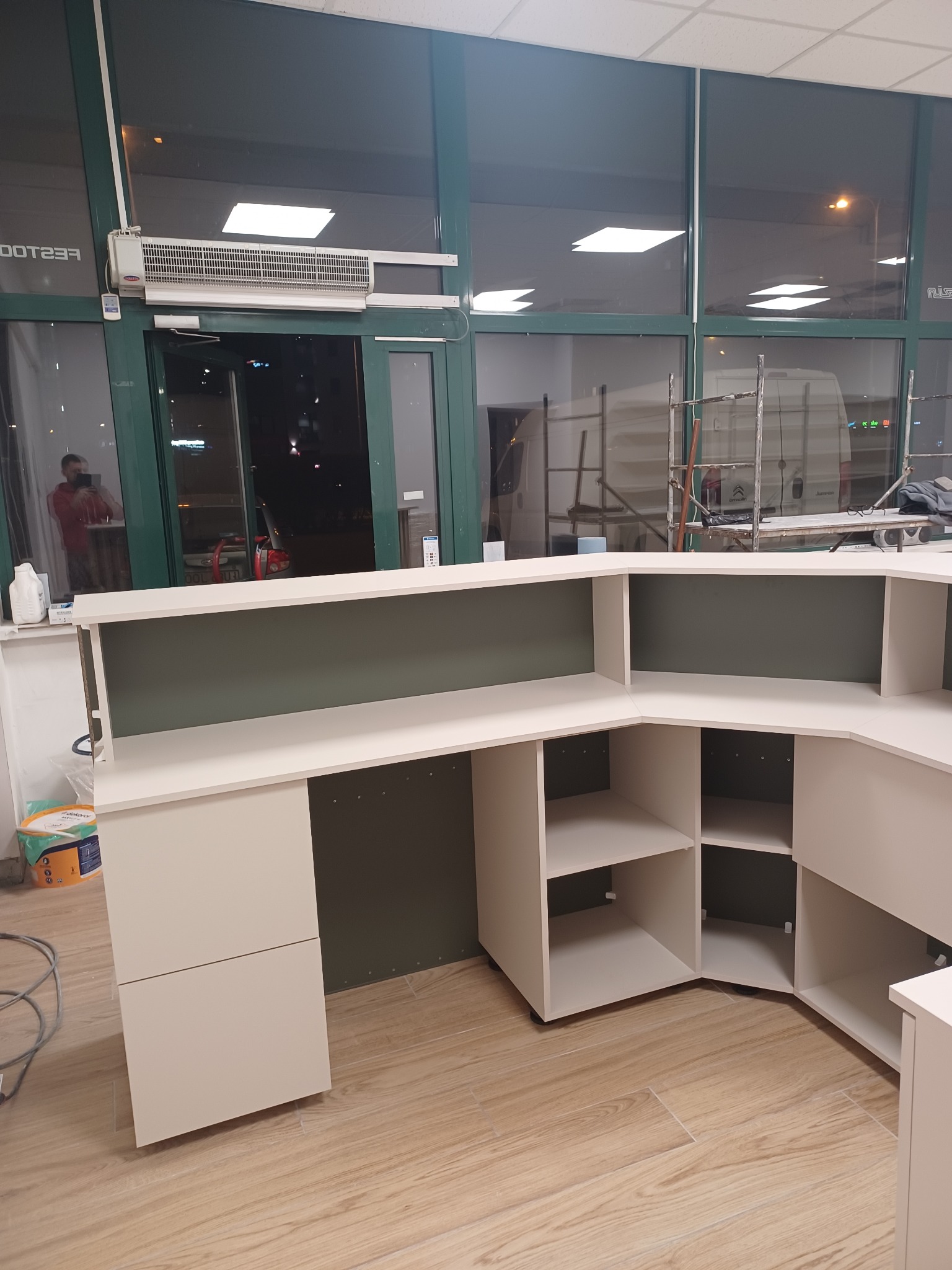 Recepcja na wymiar w trakcie montażu, widoczne puste półki i szuflady, tło stanowią okna witrynowe z widocznym rusztowaniem i odbiciem osoby.