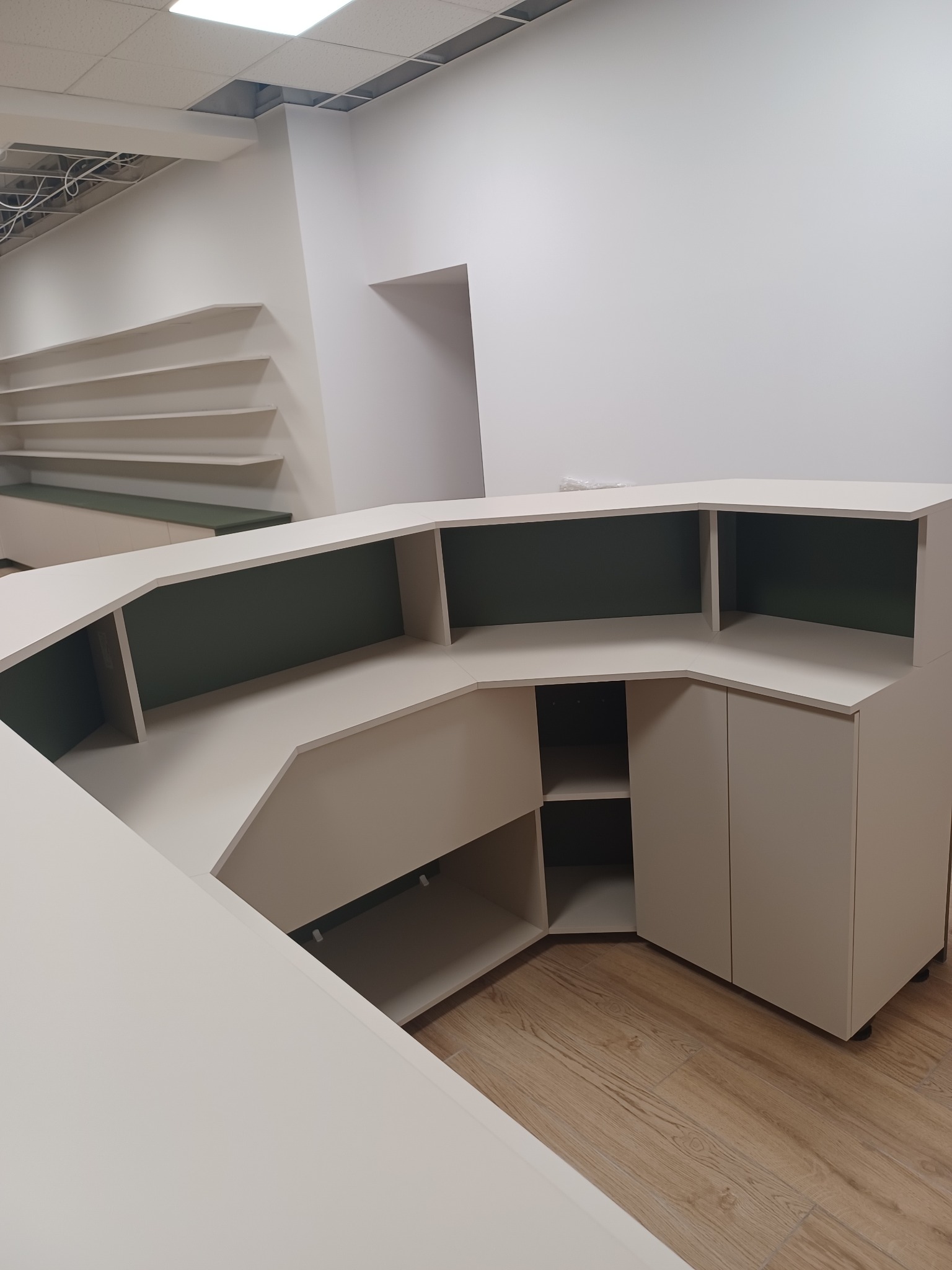 Beżowa lada recepcyjna na drewnianej podłodze w jasnym pomieszczeniu z półkami w tle, widok z perspektywy, surowy stan wykończenia.