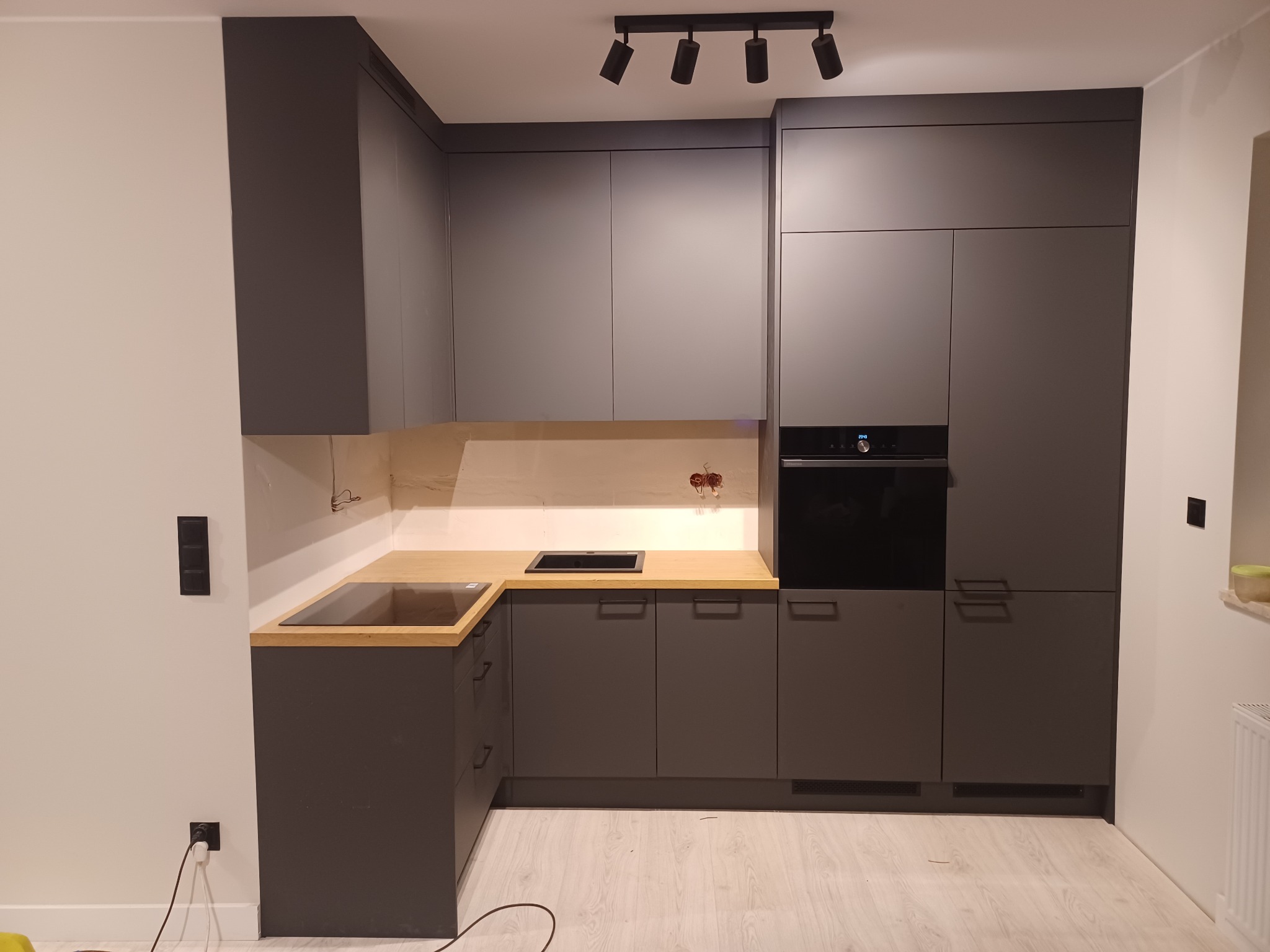 Nowoczesna, szara zabudowa kuchenna z drewnianym blatem, czarnym piekarnikiem i czarną płytą grzewczą, oświetlona reflektorami punktowymi.