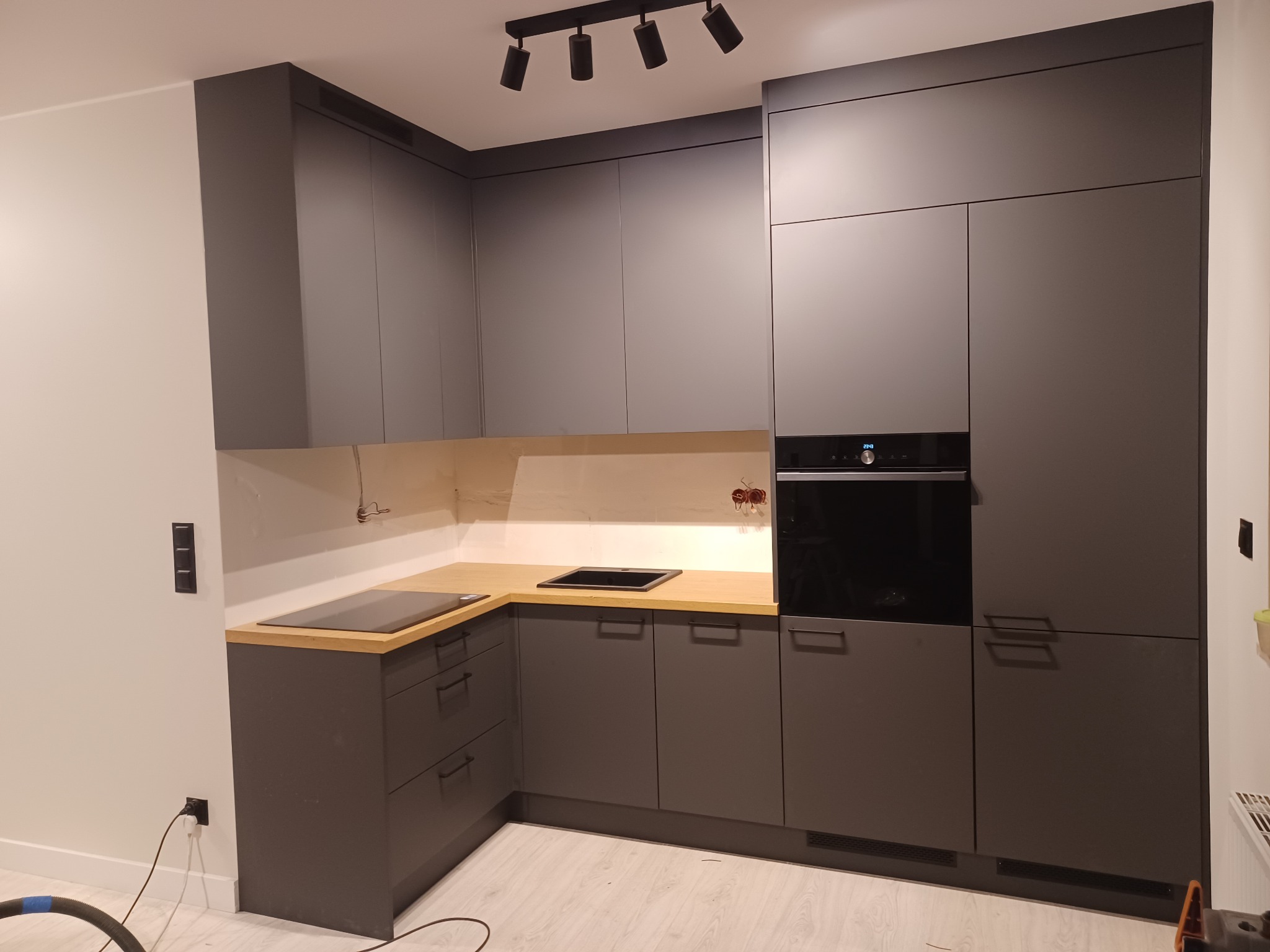 Nowoczesna, szara zabudowa kuchenna z drewnianym blatem w Rychtalu, oświetlona punktowymi lampami sufitowymi, z wbudowanym piekarnikiem i płytą indukcyjną.