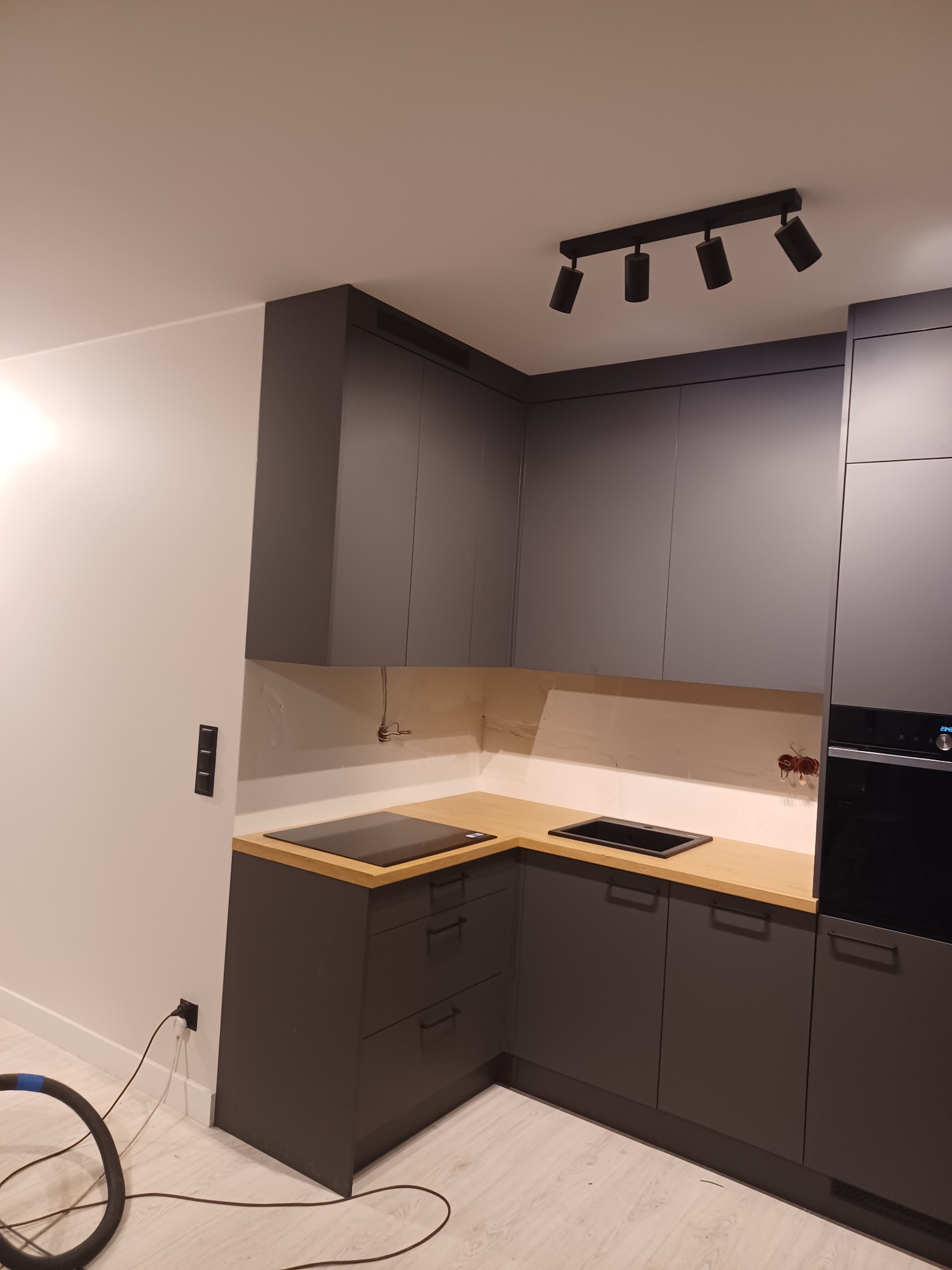 Nowoczesna kuchnia w odcieniach szarości z drewnianym blatem i czarną płytą indukcyjną, widoczne niedokończone podłączenia elektryczne i fragment odkurzacza.