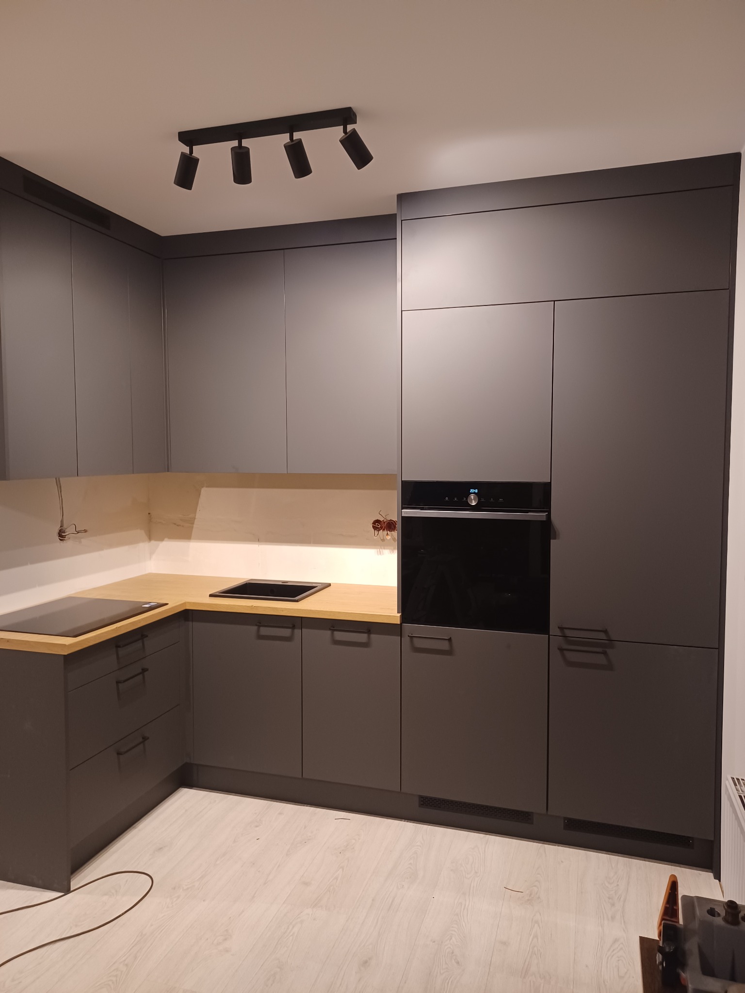 Nowoczesna kuchnia w odcieniach szarości z drewnianym blatem, oświetleniem punktowym i wbudowanym piekarnikiem, widoczne niedokończone podłączenie elektryczne.