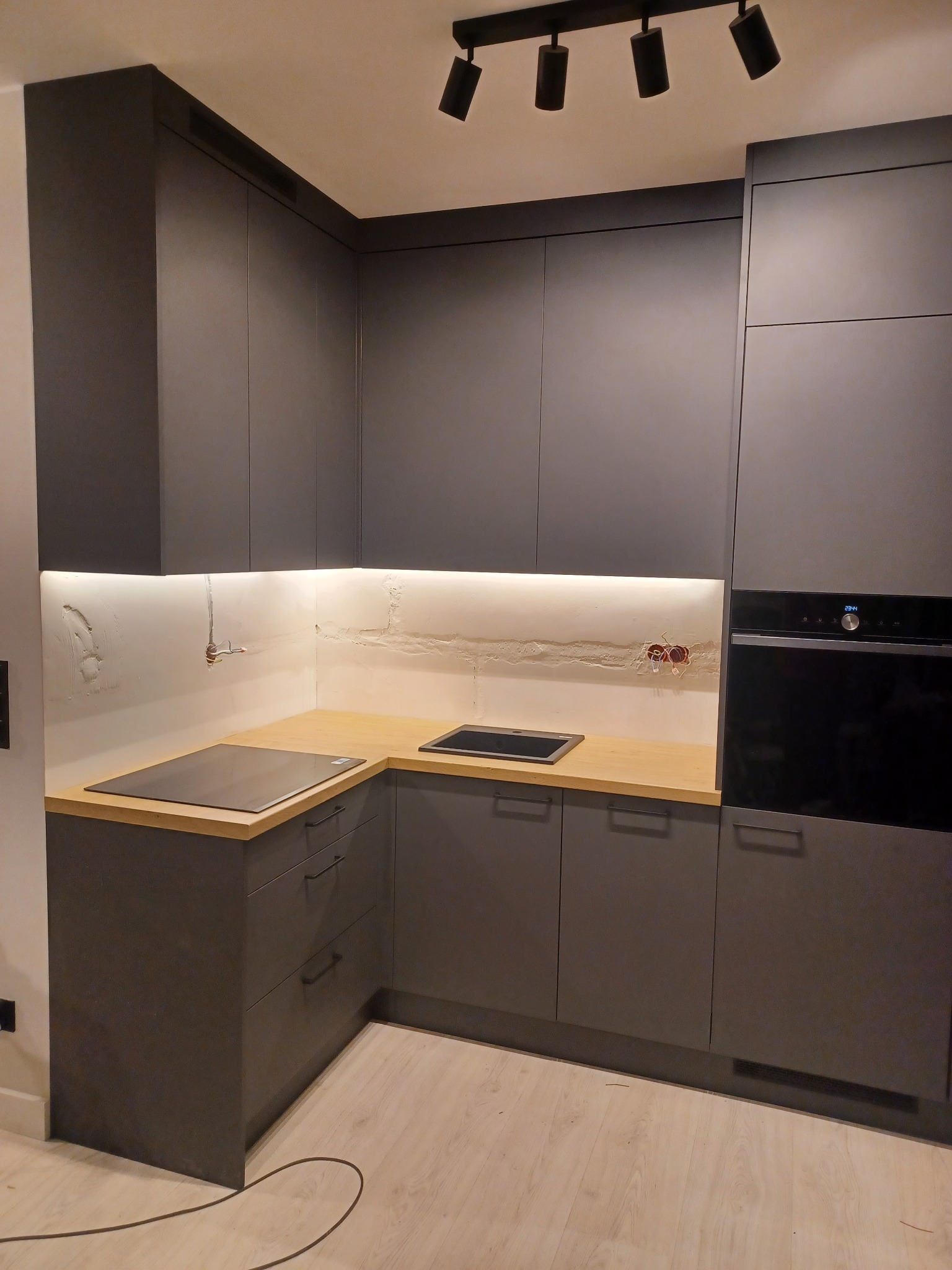 Nowoczesna, szara kuchnia z drewnianym blatem i oświetleniem LED pod szafkami, widoczne niedokończone wykończenie ściany z instalacją elektryczną.