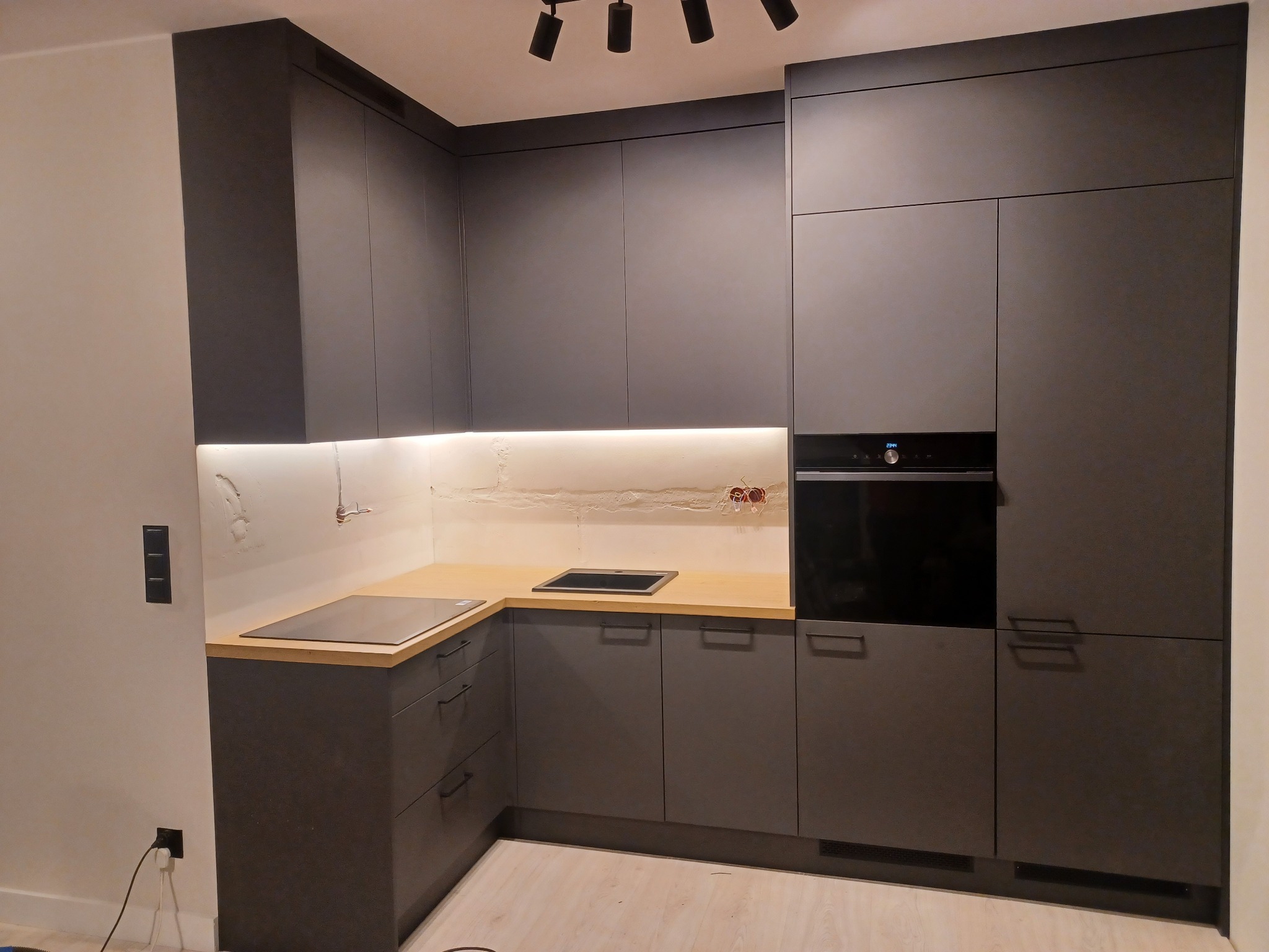 Nowoczesna, grafitowa zabudowa kuchenna z drewnianym blatem, oświetleniem LED pod szafkami wiszącymi i płytą indukcyjną, podczas montażu widoczne przewody elektryczne.