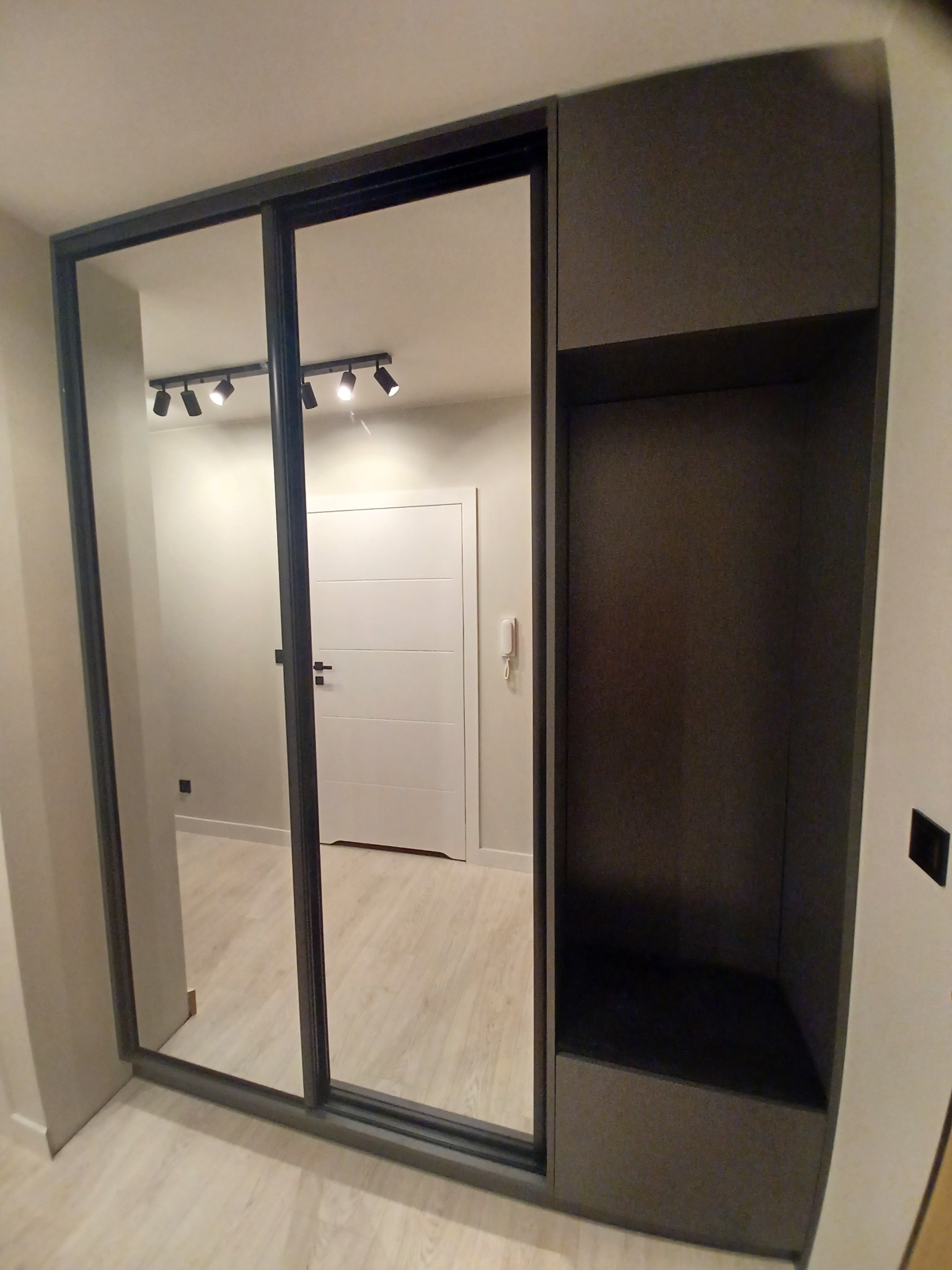Dwudrzwiowa szafa wnękowa z lustrami, czarna rama, obok otwarta półka z ciemnoszarą tapetą wewnątrz, widok na białe drzwi w tle.