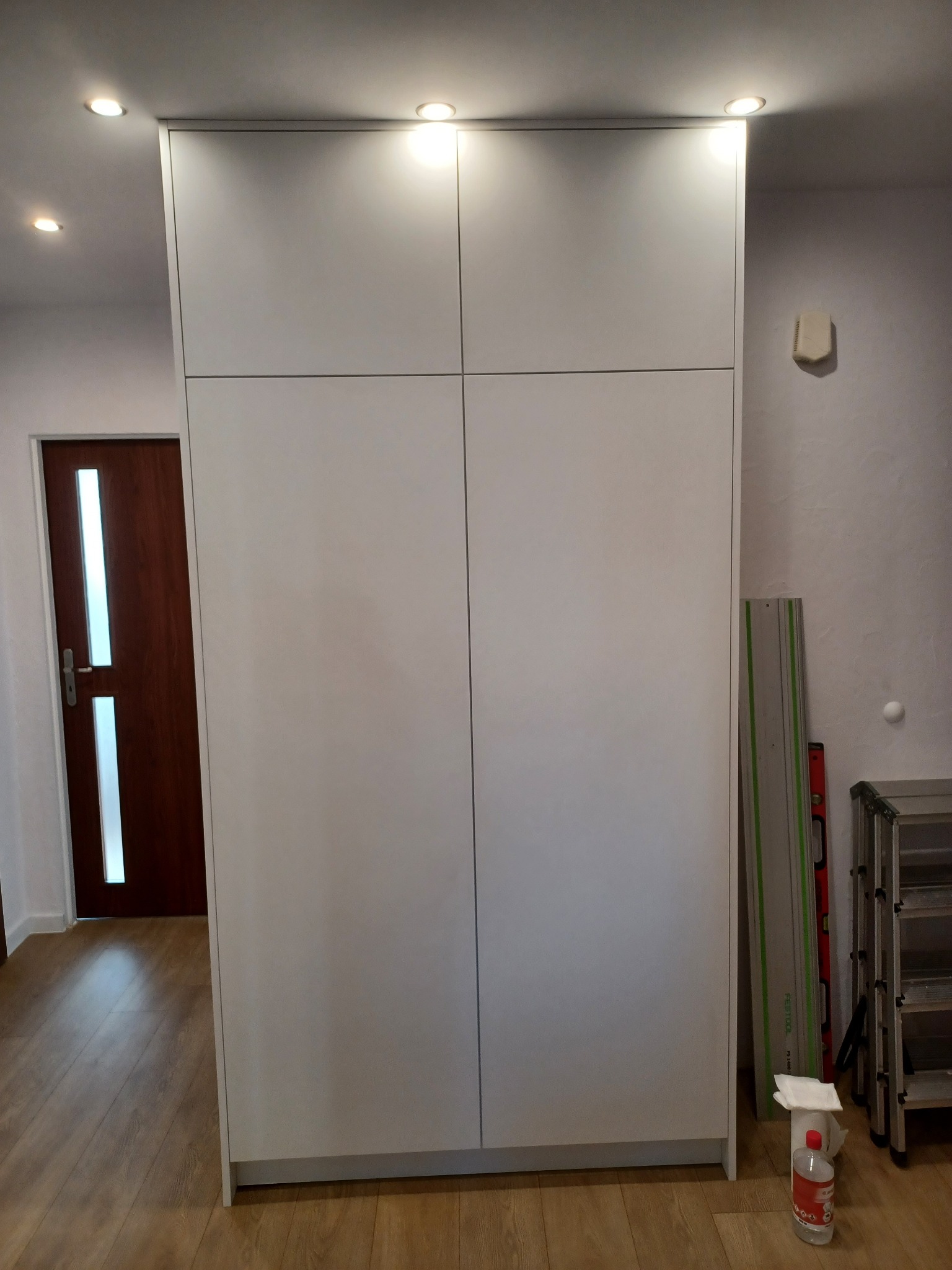 Biała szafa wnękowa z czterema drzwiami i oświetleniem punktowym w suficie, widoczne narzędzia i akcesoria montażowe obok szafy.