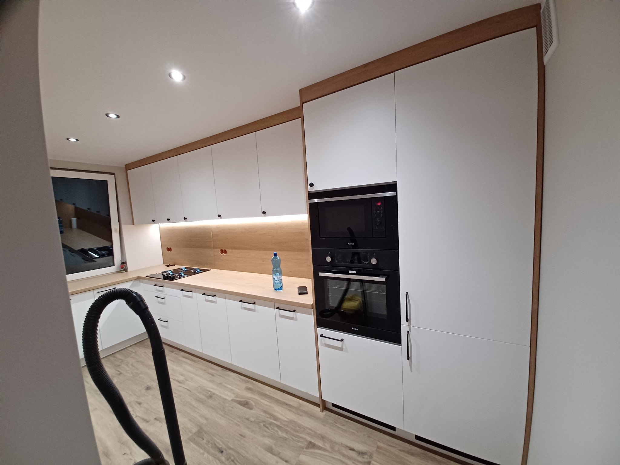 Nowoczesna kuchnia z białymi frontami i drewnianymi akcentami, widoczny piekarnik w zabudowie i fragment węża odkurzacza.