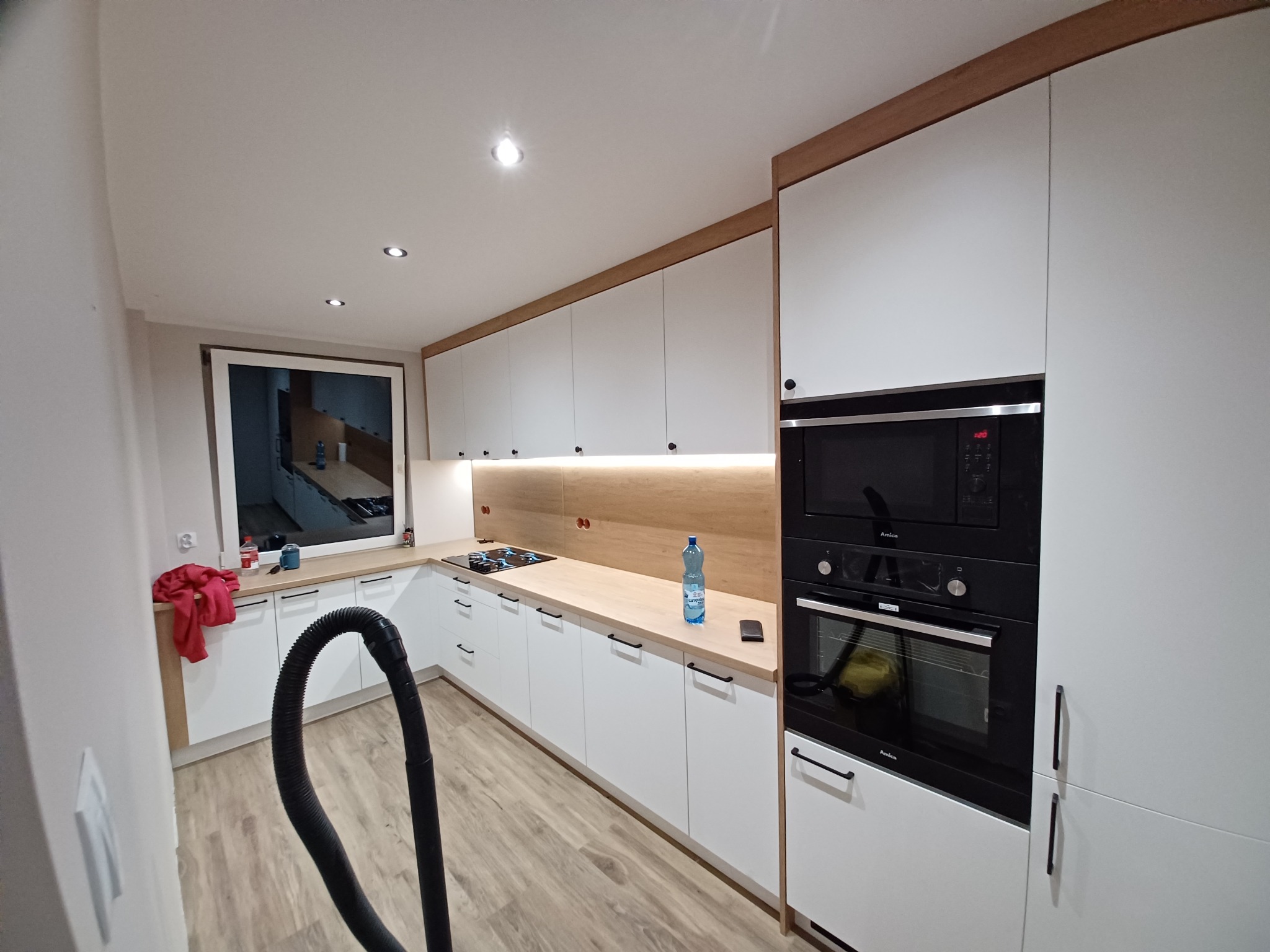 Nowoczesna kuchnia z białymi frontami, drewnianymi blatami i czarnym sprzętem AGD Amica, widoczny wąż od odkurzacza sugerujący sprzątanie po montażu.