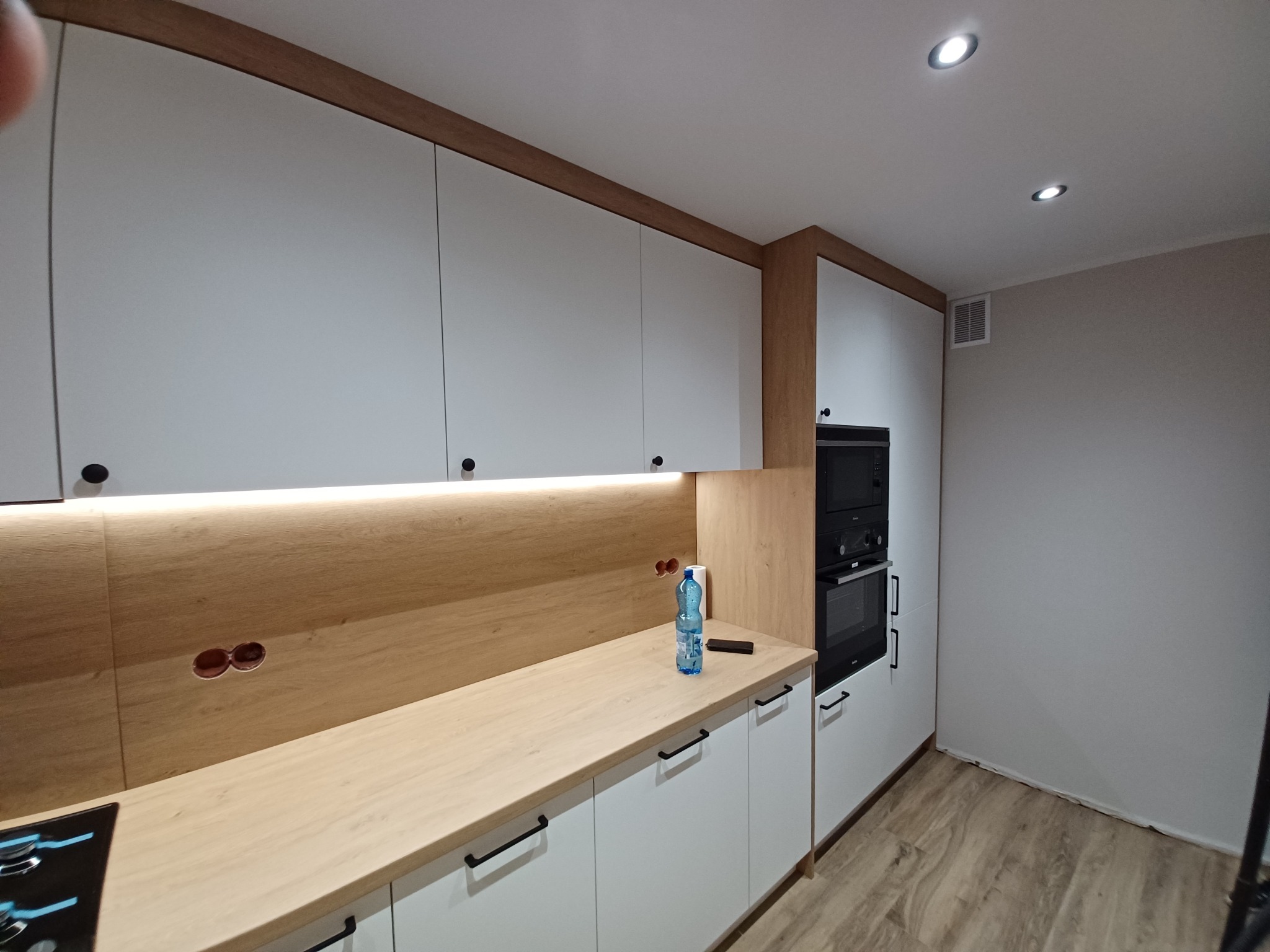 Nowoczesna kuchnia z białymi szafkami i drewnianym blatem, oświetlenie LED pod szafkami górnymi, widoczny fragment płyty indukcyjnej i butelka wody na blacie