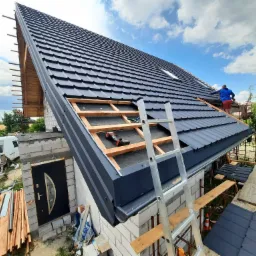 Montaż ciemnoszarej blachodachówki na nowym domu w trakcie budowy, widoczny dekarz w czerwonych spodenkach, fragment więźby dachowej, drabina oparta o budynek.