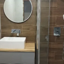 Nowoczesna łazienka z brązowymi płytkami imitującymi drewno, owalnym lustrem, białą umywalką nablatową na szarej szafce z drewnianym blatem i kabiną prysznicową typu walk-in z widocznymi...