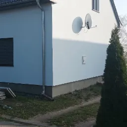 Świeżo pomalowana na biało elewacja domu jednorodzinnego z szarą dachówką, widoczne dwie anteny satelitarne i rynna spustowa, trawnik z elementami budowlanymi.