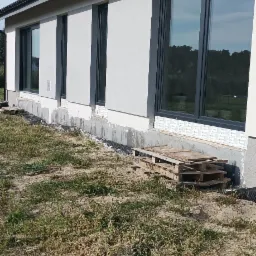 Budynek jednorodzinny z ocieploną elewacją, widoczne palety przy ścianie, etap prac wykończeniowych.