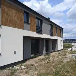 Dwukondygnacyjny segment bliźniaka z szarą i białą elewacją oraz drewnopodobnymi panelami na piętrze, widok z poziomu gruntu, w tle błękitne niebo z chmurami.