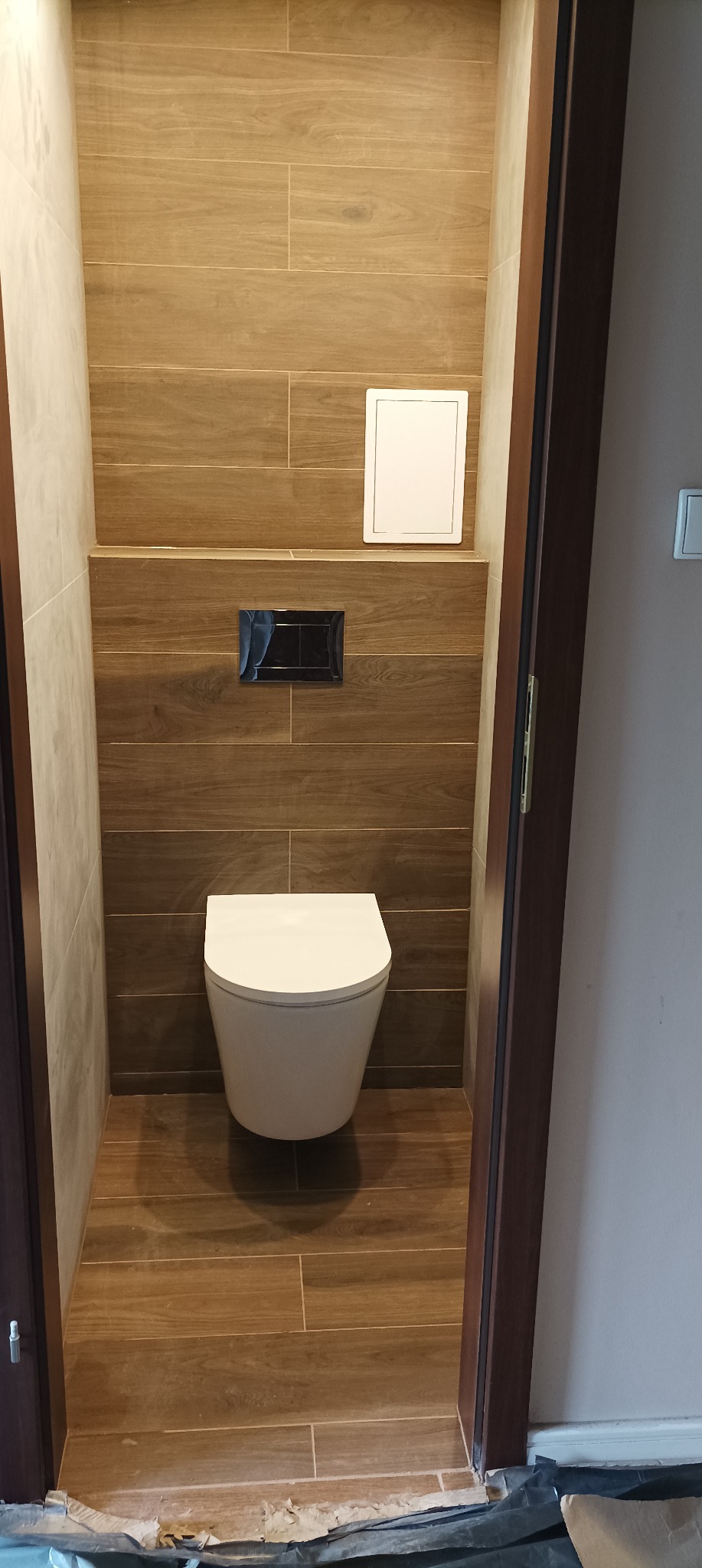 Nowoczesna toaleta podwieszana w małej łazience, brązowe płytki imitujące drewno na ścianach i podłodze, czarny panel sterujący spłuczką, biała klapa rewizyjna nad toaletą, widoczne niedociągnięcia...