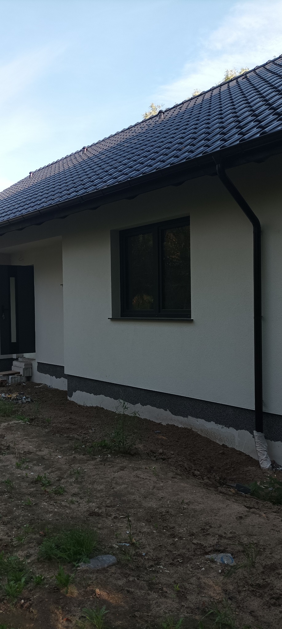 Nowoczesny dom z jasną elewacją, ciemną stolarką okienną i grafitowym dachem z dachówki ceramicznej, widok na fragment ściany bocznej z oknem i rynną, teren wokół domu jeszcze niezagospodarowany.