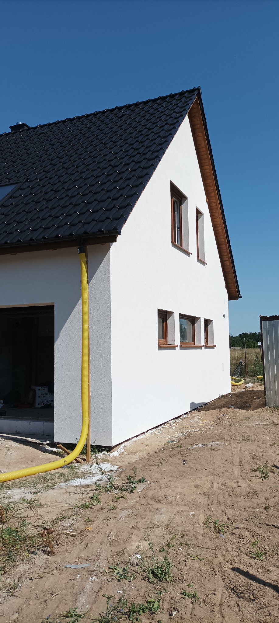 Nowo otynkowany, biały dom jednorodzinny z ciemną dachówką, widoczny żółty wąż wentylacyjny i teren wokół w trakcie zagospodarowywania.