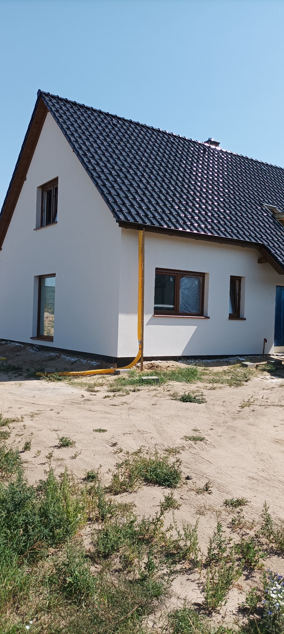 Nowo otynkowany dom jednorodzinny z brązowymi oknami i ciemną dachówką, widoczny żółty system odprowadzania wody deszczowej, teren wokół domu częściowo zagospodarowany.