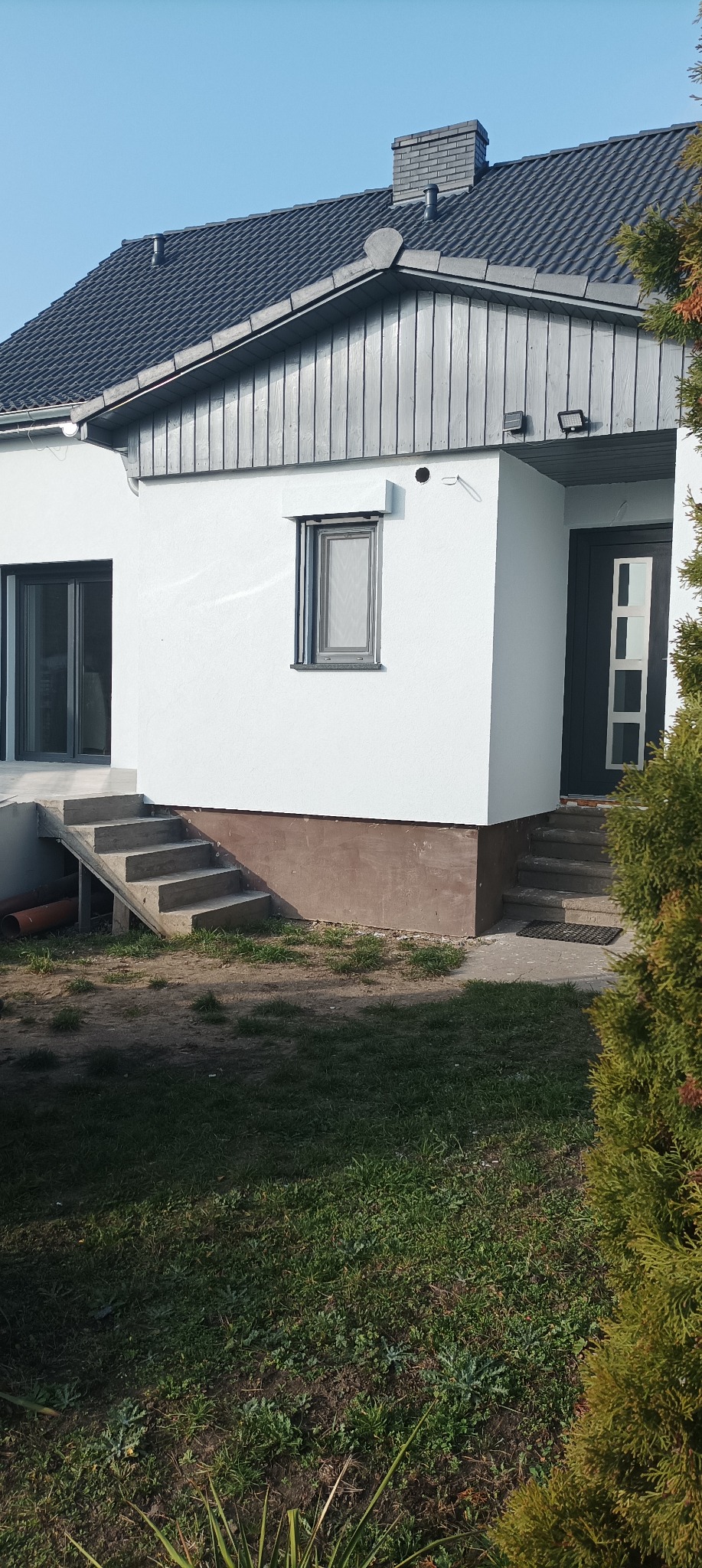 Nowoczesny dom z jasną elewacją, ciemnoszarą stolarką okienną i drzwiami wejściowymi, oraz schodami z betonu, widziany z perspektywy ogrodu.