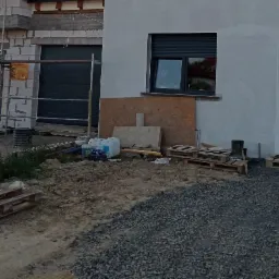 Budynek w trakcie budowy: widoczna ściana z oknem z roletą, brama garażowa w surowym stanie, rusztowania, płyty OSB, palety i pojemniki z materiałami budowlanymi na placu budowy.