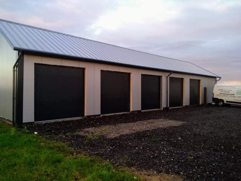 Budynek magazynowy z czterema czarnymi bramami garażowymi rolowanymi, srebrny dach, szara elewacja, zaparkowany biały samochód dostawczy z logo firmy w tle.