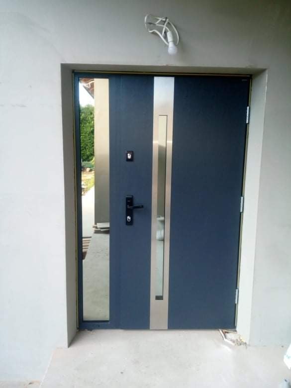 Nowoczesne, niebieskie drzwi wejściowe z pionowym, metalowym panelem i bocznym przeszkleniem, zamontowane w surowym, szarym otworze ściennym, z widoczną żarówką bez klosza na górze.