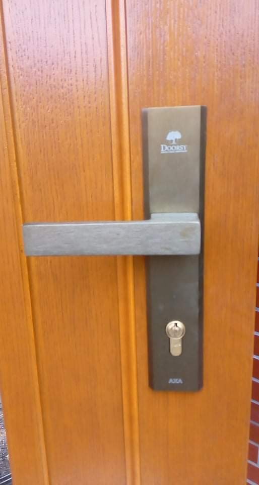 Zamknięte drewniane drzwi z nowoczesną, prostokątną klamką i zamkiem marki Doorsy oraz widocznym logo AXA w dolnej części zamka.