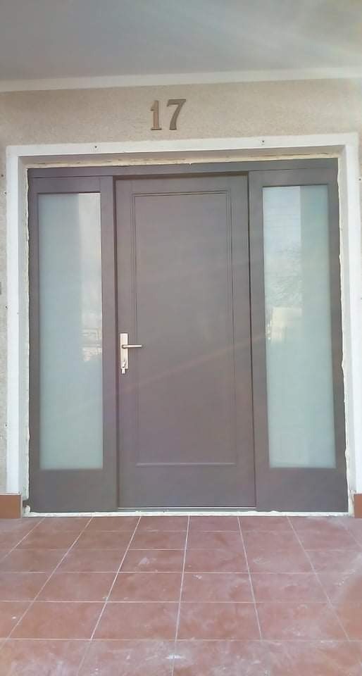 Szare drzwi wejściowe z bocznymi szybami i numerem domu '17' nad nimi, widoczne z zewnątrz na tle beżowej elewacji i brązowych płytek podłogowych.