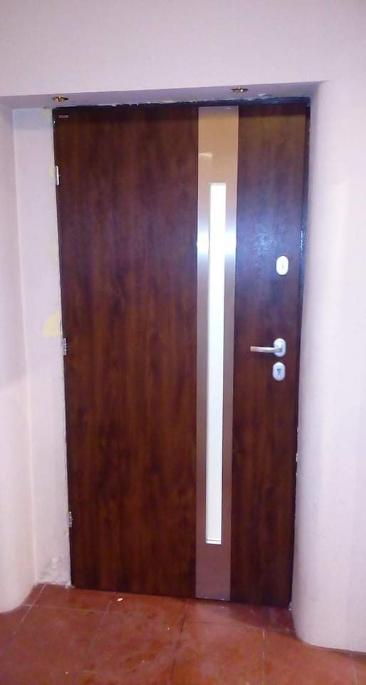 Brązowe drzwi wejściowe z pionowym srebrnym paskiem i wizjerem, zamontowane w białej futrynie wewnątrz budynku.