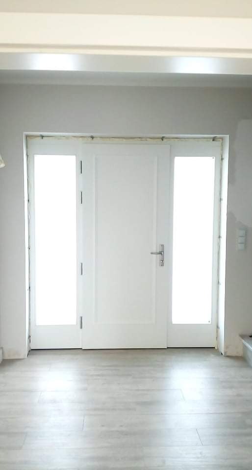 Białe drzwi wejściowe z bocznymi panelami szklanymi, widoczne ślady montażu na ścianie i podłodze, surowe wykończenie wnętrza.