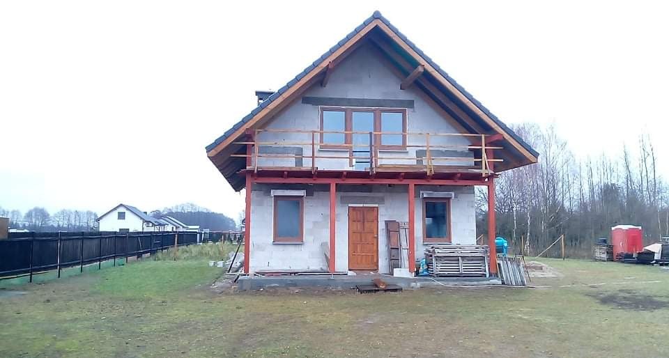 Dom w trakcie budowy z widocznymi oknami z roletami zewnętrznymi, drewnianymi drzwiami wejściowymi i balkonem z drewnianą balustradą, widoczne rusztowania i materiały budowlane.