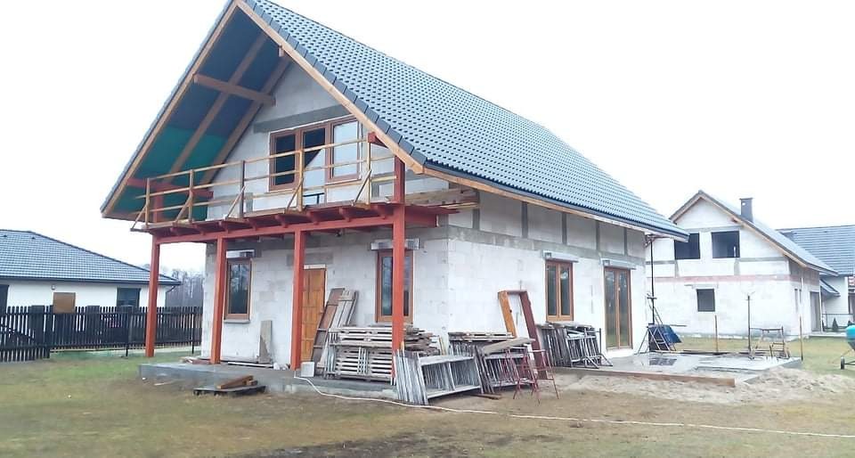 Dom w stanie surowym otwartym z widocznymi elementami konstrukcyjnymi balkonu, oknami bez szyb, paletami i ramami okiennymi przed budynkiem.