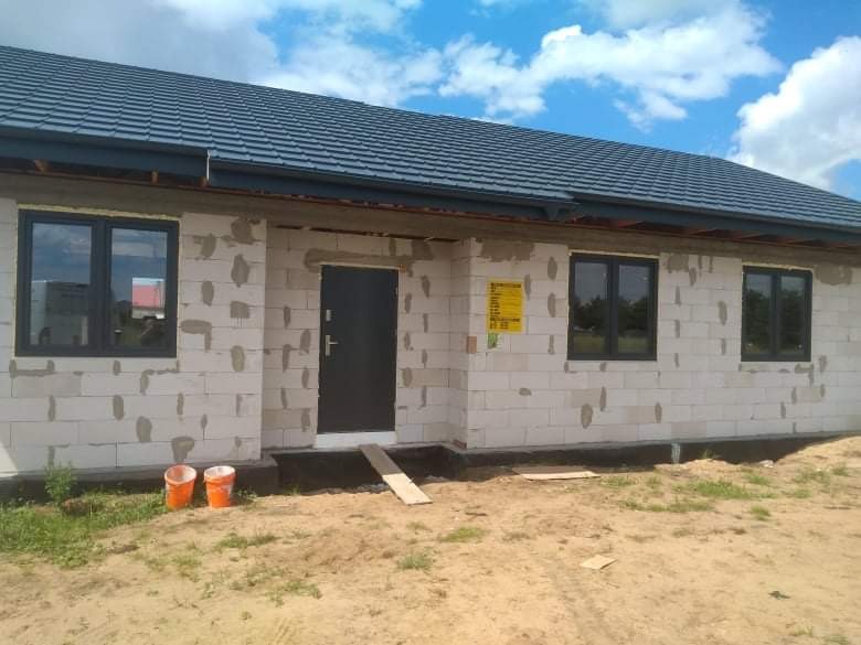 Surowy budynek w trakcie budowy z szarymi oknami i drzwiami wejściowymi, widoczne dwa pomarańczowe wiadra na ziemi i deska przed wejściem.