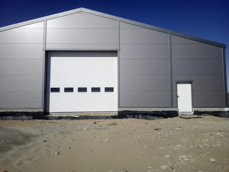Szary budynek przemysłowy z białą segmentową bramą garażową i drzwiami wejściowymi, widoczne czarne uszczelki przy fundamencie.