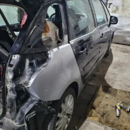 Ciemny samochód w trakcie naprawy blacharskiej; otwarte drzwi bagażnika, widoczne uszkodzenia karoserii, fragmenty zabezpieczone taśmą, nowa, surowa blacha w miejscu uszkodzeń, narzędzia...