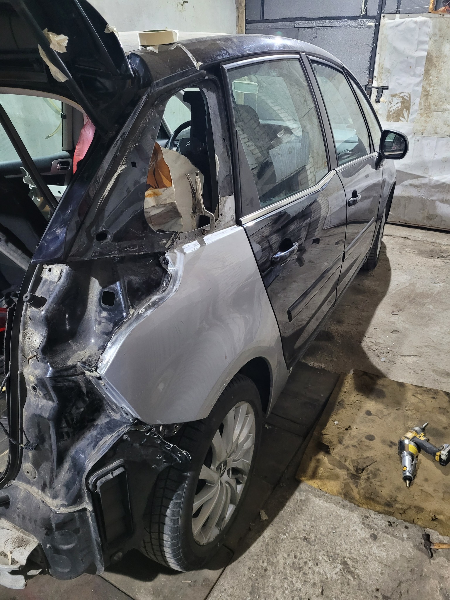 Ciemny samochód w trakcie naprawy blacharskiej; otwarte drzwi bagażnika, widoczne uszkodzenia karoserii, fragmenty zabezpieczone taśmą, nowa, surowa blacha w miejscu uszkodzeń, narzędzia...