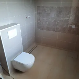 Zabudowany, podwieszany sedes w łazience wykończonej beżowymi płytkami; na ścianie płytki imitujące marmur, widoczne przyłącza wodne.