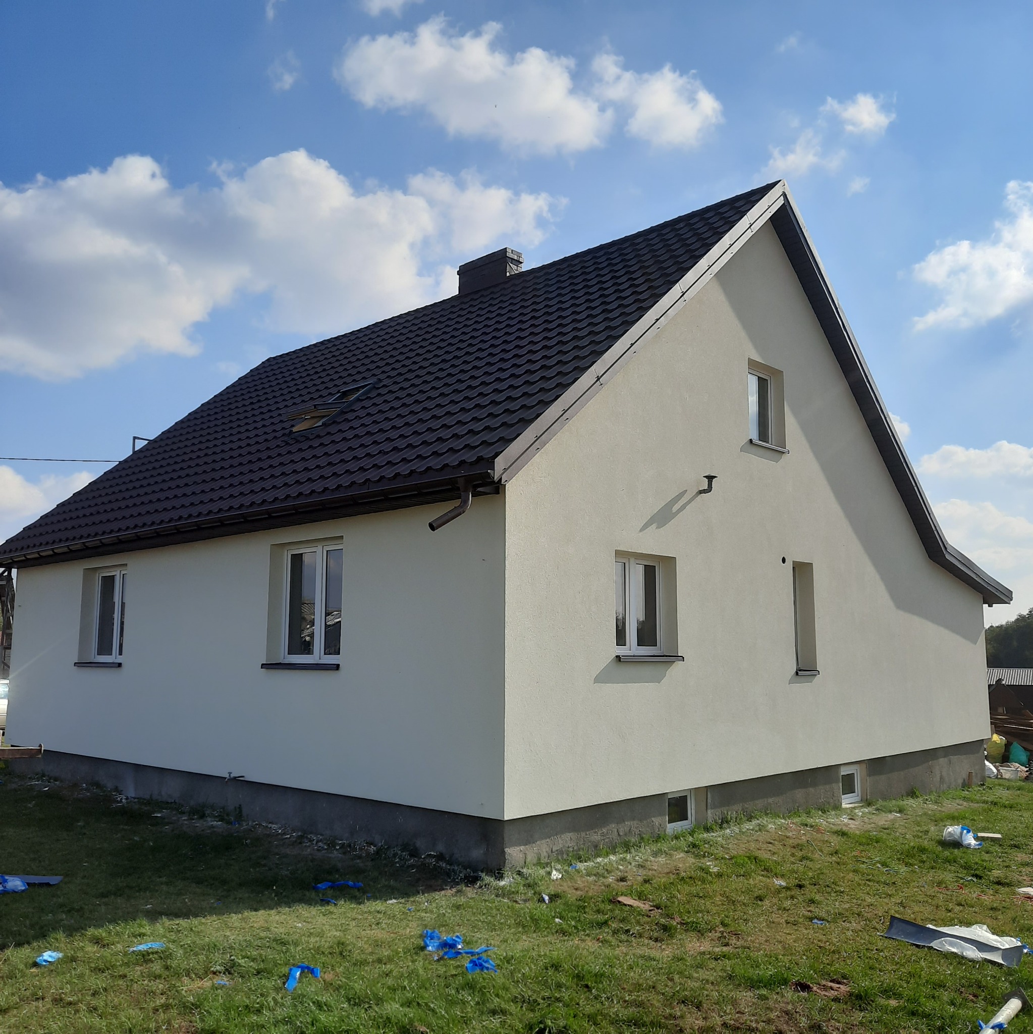 Świeżo otynkowany dom jednorodzinny z ciemną dachówką, widok z zewnątrz, na trawie leżą resztki folii malarskiej, słoneczny dzień.