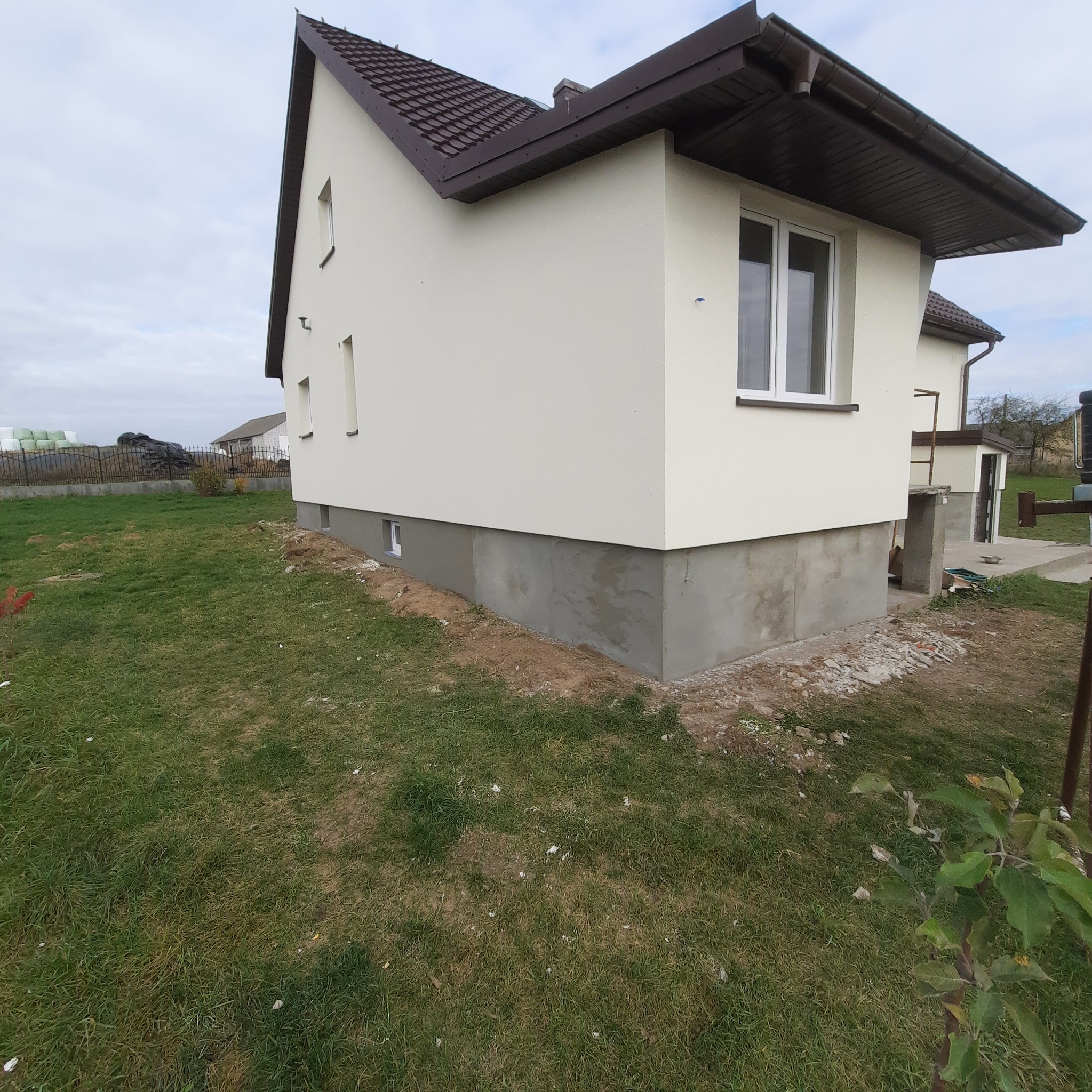 Świeżo otynkowana elewacja domu jednorodzinnego z widocznym fragmentem podmurówki w surowym stanie, otoczona trawnikiem i elementami krajobrazu.