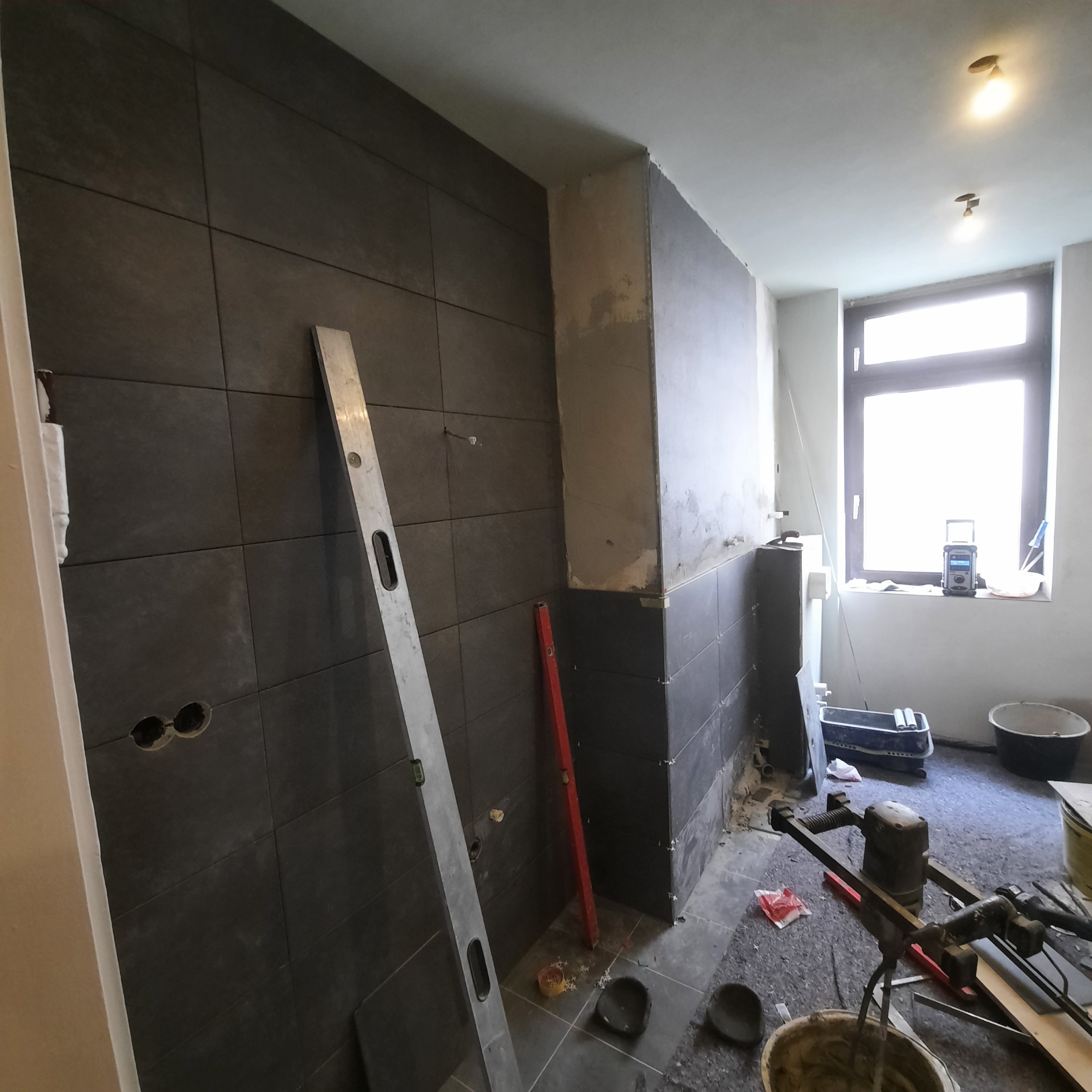 Ciemne płytki na ścianie łazienki w trakcie montażu, widoczny poziomica, mieszadło do zaprawy, okno z radiem i częściowo położone płytki.