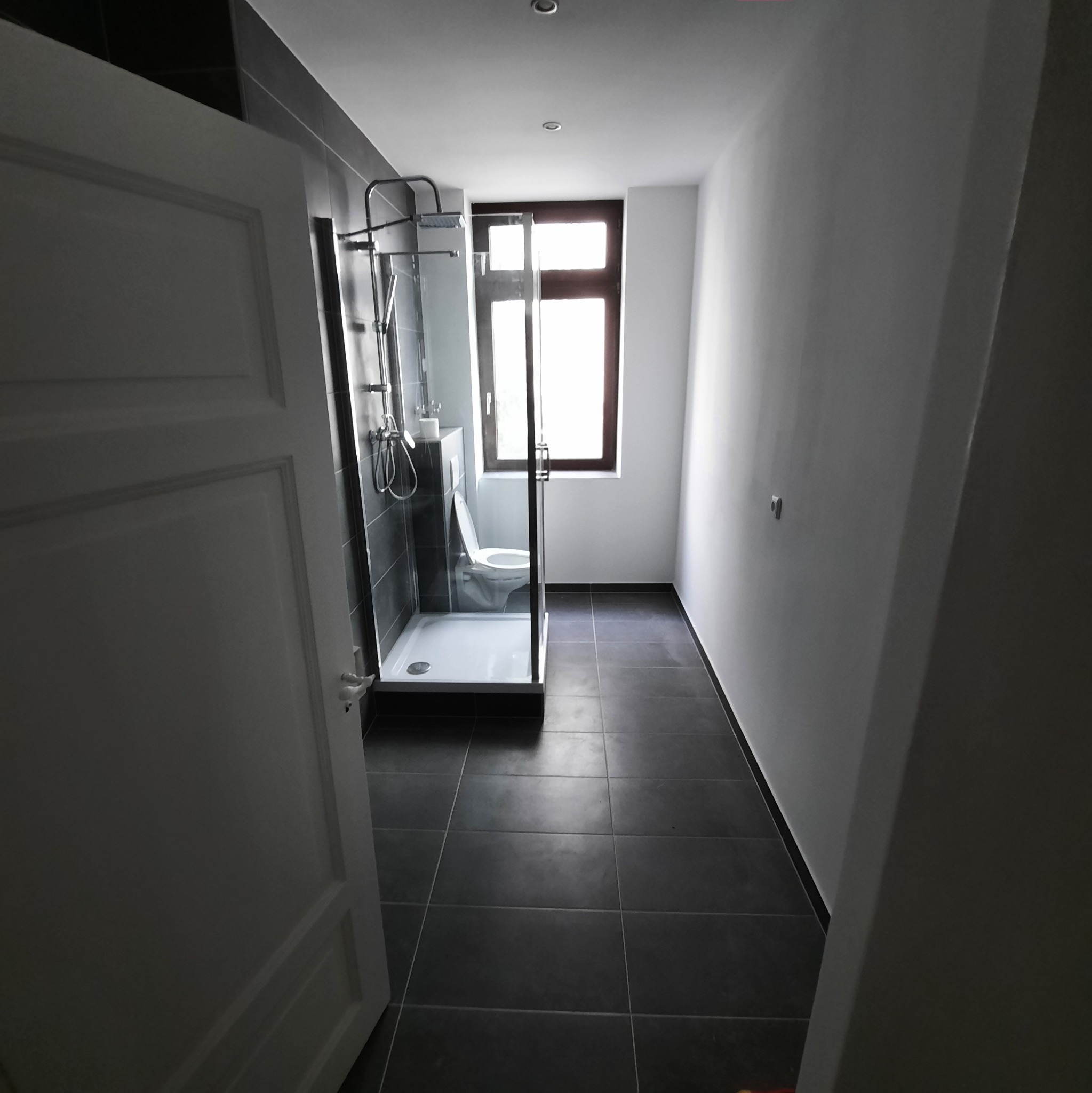 Wyremontowana łazienka z kabiną prysznicową typu walk-in, czarnymi płytkami podłogowymi i ściennymi, białymi ścianami oraz uchylonymi białymi drzwiami wejściowymi.