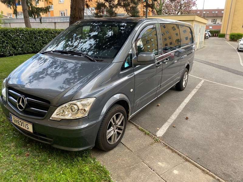 Szary Mercedes Vito zaparkowany na parkingu, widoczny z boku, z zielonym trawnikiem po lewej stronie i budynkami w tle.
