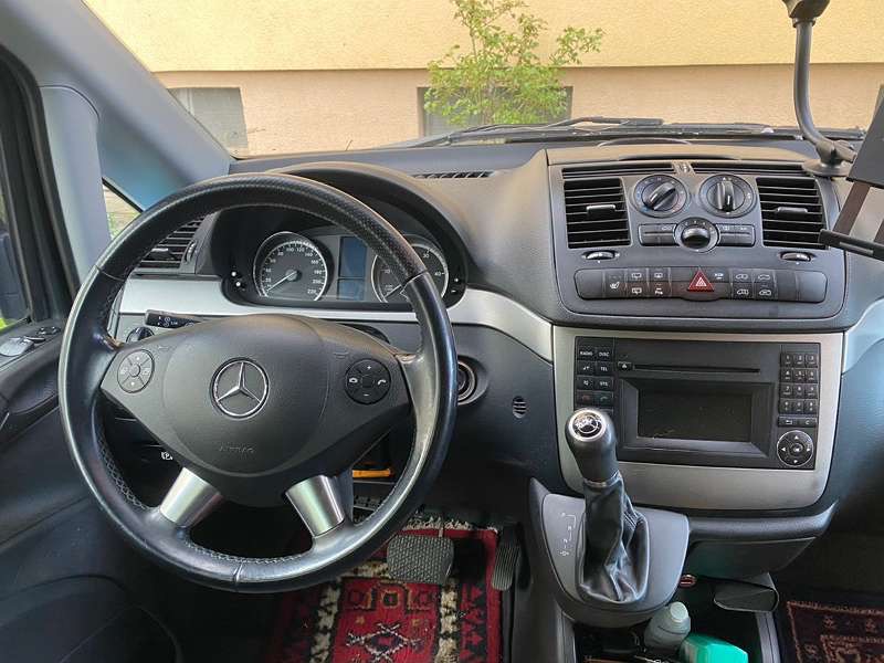 Wnętrze samochodu dostawczego Mercedes-Benz z kierownicą po lewej stronie, widoczny kokpit, radio, dźwignia zmiany biegów i dywanik z wzorem geometrycznym.
