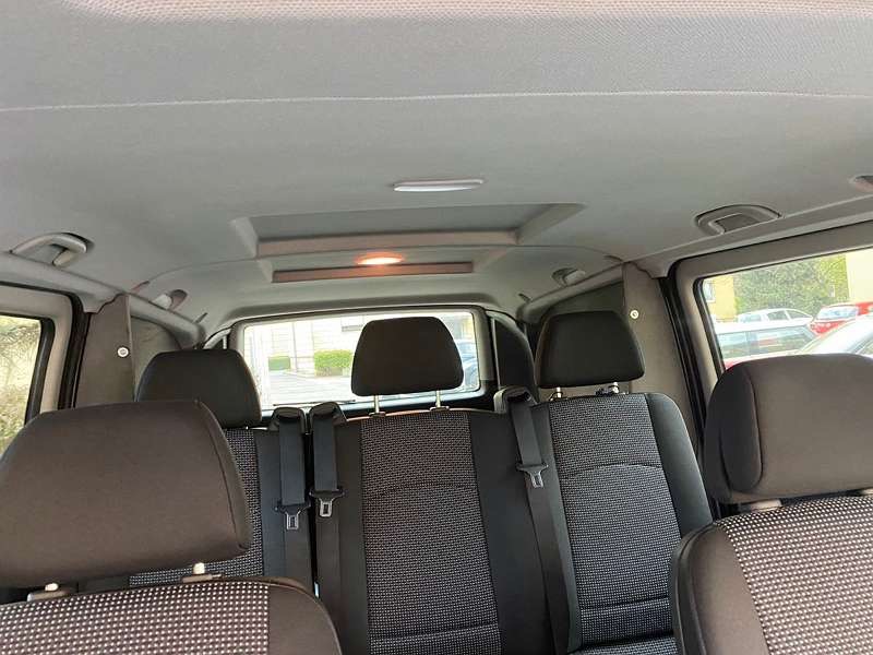 Wnętrze busa z widocznymi tylnymi siedzeniami z szarej tapicerki w geometryczny wzór, zagłówkami i pasami bezpieczeństwa. Widoczne okna boczne oraz oświetlenie sufitowe.